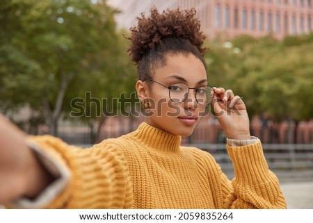 Serious female model with combed curly hair wears round spectacles yellow knitted sweater stretches arm with unrecognizable device takes photo of herself while strolling at street during leisure time