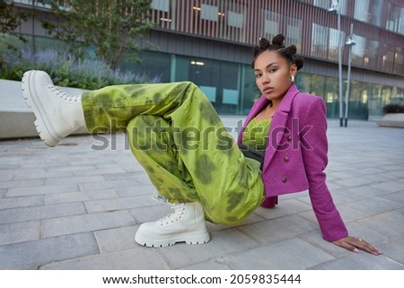 Horizontal shot of stylish teenage girl wears fashionable cloths and white boots has bright makeup poses against urban building poses for making photo practices breakdancing looks seriously. Royalty-Free Stock Photo #2059835444