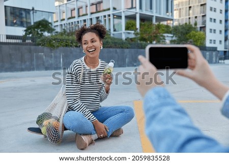 Happy millennial girl laughs positively sits crossed legs on skateboard carries net bag with fruits holds bottle of water looks cheerfully away her unrecognizable friend takes photo on smartphone