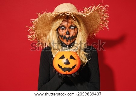 Young scary woman with Halloween makeup mask wears straw hat black scarecrow costume hold pumpkin jack-o-lantern isolated on plain red background studio portrait. Celebration holiday party concept.
