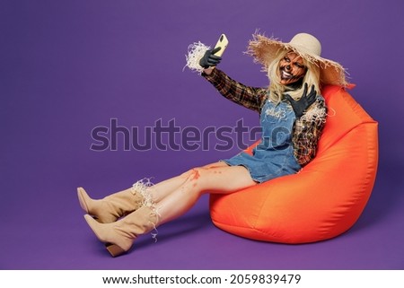 Full body young woman with Halloween mask in hat scarecrow costume sit in orange bag chair do selfie shot mobile cell phone say arrr isolated on plain dark purple background studio Celebration concept
