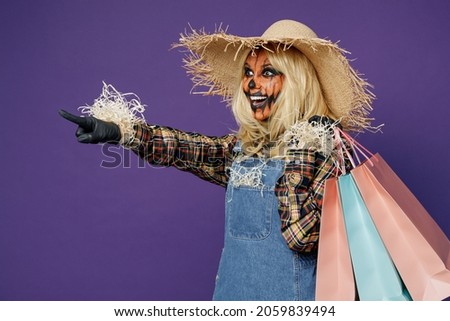 Side view young woman with Halloween makeup in hat scarecrow costume hold package bags, purchases after shopping point finger aside isolated on plain dark purple background studio Celebration concept