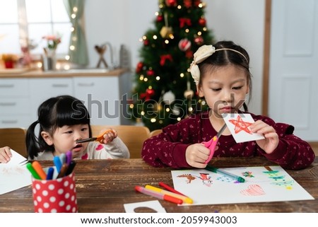 innocent asian baby girl is holding and looking at scissors with curiosity while doing paper craft with her older sister in Christmas home interior on background