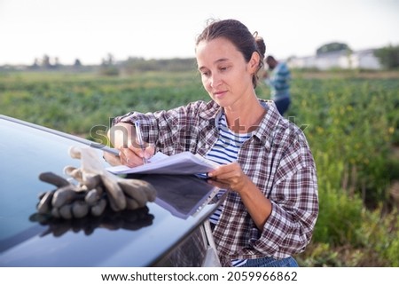 Confident female farmer standing outdoors near car on background with farm field on fall day, signing some papers