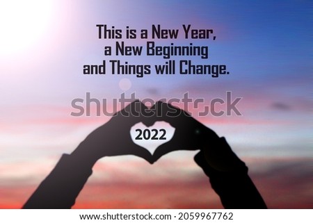 New year inspirational motivational quote - This is a new year a beginning, things will change. Silhouette of a person holding a heart shaped hands with 2022 number inside. On sunset sky background. Royalty-Free Stock Photo #2059967762