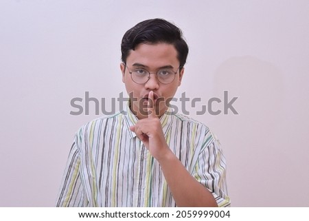 Portrait of handsome Asian man in casual stripe shirt with glasses making shushing gesture, asking to be quiet, silence concept. Isolated image on gray background