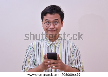 Portrait of funny Asian man in casual stripe shirt with glasses holding a mobile phone with two hands and looking at camera. Isolated image on gray background Royalty-Free Stock Photo #2060006699