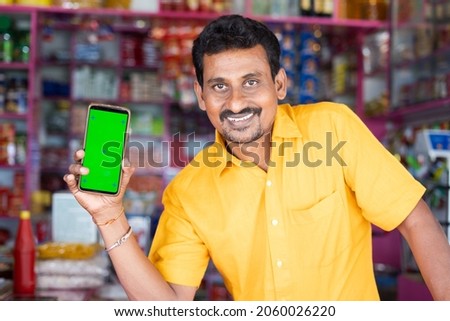 Smiling Merchant at groceries store hold mobile with green screen mock up by looking at camera - concept of Technology, advertisement, online booking and e-commerce. Royalty-Free Stock Photo #2060026220