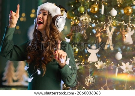 Christmas time. happy stylish woman with Santa hat in green dress listening to the music with headphones and dancing near Christmas tree at modern home. Royalty-Free Stock Photo #2060036063