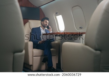 Handsome pensive middle eastern businessman talking on mobile phone, holding financial report working with documents sitting in plane. Confident entrepreneur flying private jet, successful business Royalty-Free Stock Photo #2060036729