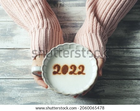 Number 2022 on frothy surface of cappuccino served in pastel pink cup holding by female hands over grey painted wood planks table. Holidays food art theme Happy New Year 2022, New year new you. Royalty-Free Stock Photo #2060065478