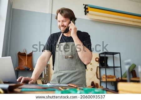 Low angle portrait of smiling male artisan using laptop and speaking by smartphone while working in tanners workshop, copy space