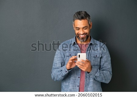 Mid adult multiethnic man texting phone message on smart phone isolated on grey background. Smiling middle eastern man using smartphone on gray wall. Happy mixed race guy using app on mobile phone. Royalty-Free Stock Photo #2060102015