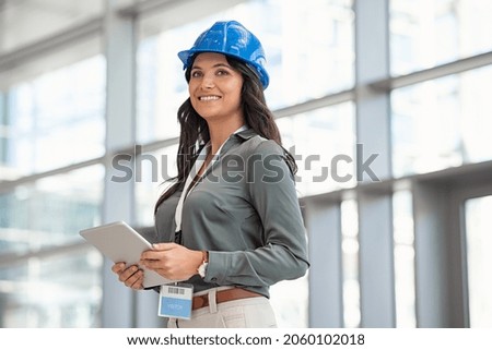 Mid adult woman architect wearing hardhat at construction site while working on digital tablet. Supervisor wearing safety helmet while working in a building site. Successful and proud inspector. Royalty-Free Stock Photo #2060102018