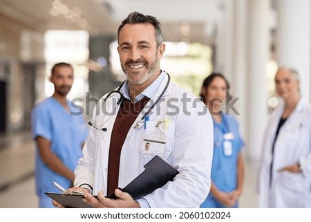 Portrait of mature doctor wearing lab coat and stethoscope reading patient case file in hospital. Successful head physician standing in hospital hallway and looking at camera with his medical team. Royalty-Free Stock Photo #2060102024