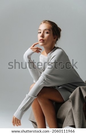Looking at you teenage girl sitting on an armchair, leaning forward touching her cheek. She's wearing warm longsleeve covering her body down to her thighs with bare legs showing under.