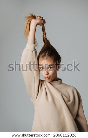 Comical grimacing teenage girl pulling her hair up, making kissing lips. Looking at the camera. Against grey background. In a warm beige longsleeve.
