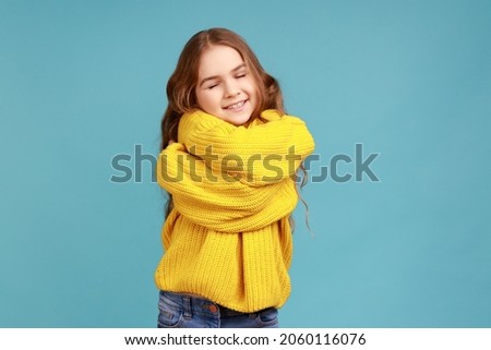 Portrait of charming little girl embracing herself and smiling from happiness, positive self-esteem, wearing yellow casual style sweater. Indoor studio shot isolated on blue background. Royalty-Free Stock Photo #2060116076