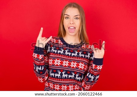 Young beautiful caucasian girl wearing christmas sweaters on red background making rock hand gesture and showing tongue