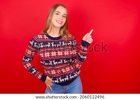 Young beautiful caucasian girl wearing christmas sweaters on red background looking at camera indicating finger empty space sales