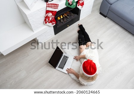 Cheerful woman making video call on Christmas day and greeting somebody, looks happy, clenching fist like winner, looks at screen of device, posing indoor near fireplace