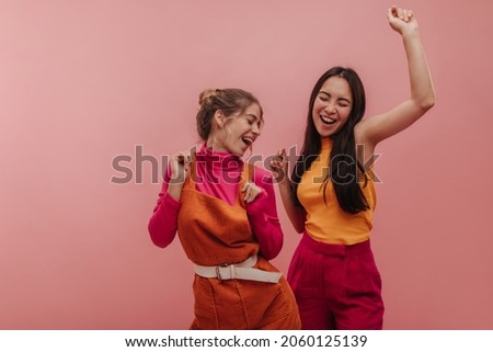 Studio portrait of young asian and european woman happily singing and dancing in pink studio. Slim brunette and blonde with closed eyes in autumn casual clothes are having fun.