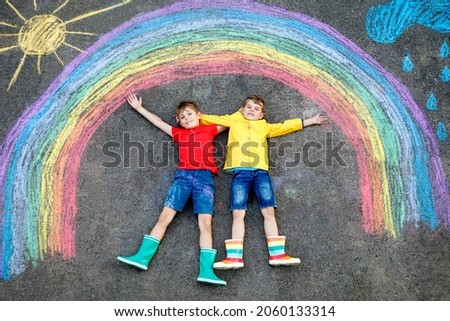 Two happy school boys having fun with with rainbow picture drawing with colorful chalks on asphalt. Siblings, twins and best friends painting on ground together. Sign against corona virus.