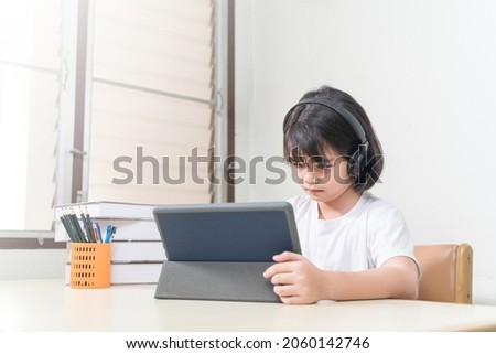 Cheerful Asian little girl student wears headphone, writes on digital tablet to study at home. Education Concept Stock Photo