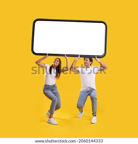 Millennial couple holding huge heavy smartphone with white empty screen on yellow orange studio background. Mock up for mobile app or new website advertising, great offer. Cellphone display template Royalty-Free Stock Photo #2060144333