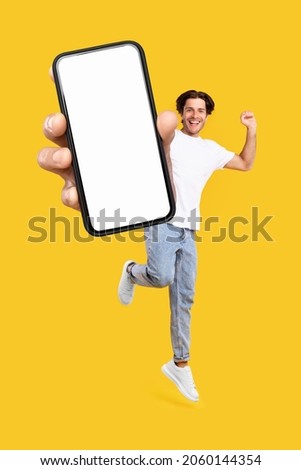 Yes, Cool Mobile Offer Ad. Joyful young casual guy showing big empty cell phone screen jumping up in the air on orange studio background, creative collage. Happy man recommend website, free copy space Royalty-Free Stock Photo #2060144354