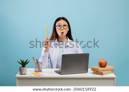 I have great idea. Emotional asian lady pointing pencil up, thinking and finding inspiration, sitting at desk with laptop over blue background, studio shot Royalty-Free Stock Photo #2060158922