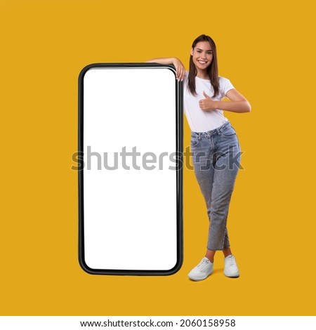 Cool Mobile Offer. Happy casual lady leaning on huge cellphone with empty white screen, showing thumb up, recommending great new app or website for cell phone, offering space for your ad, mock up