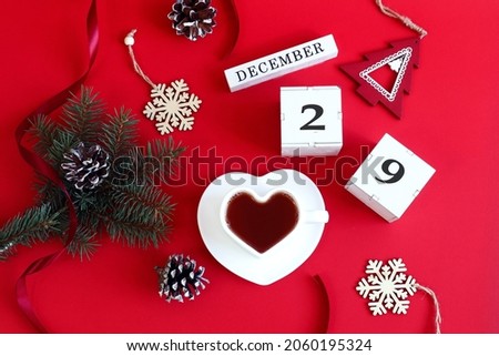 Calendar for December 29: the name of the month in English, cubes with the number 29, a cup of tea in the shape of a heart, a fir branch, cones, Christmas decor on a red background, top view