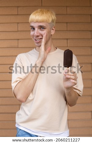 Junk food, summer, sweets and people concept - A whispering Caucasian man dressed in a beige T-shirt and holding a chocolate ice cream cone on a stick looking at the camera outside on a brick wall 
