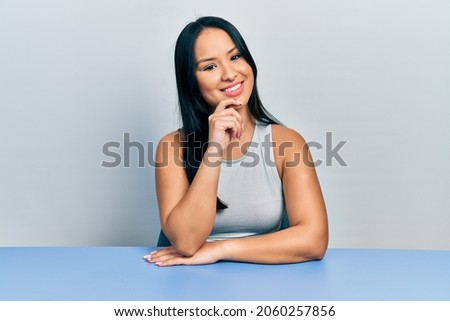 Beautiful hispanic woman with nose piercing sitting on the table looking confident at the camera with smile with crossed arms and hand raised on chin. thinking positive. 