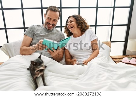 Middle age hispanic couple reading book and using smartphone. Lying on the bed with dog at home.