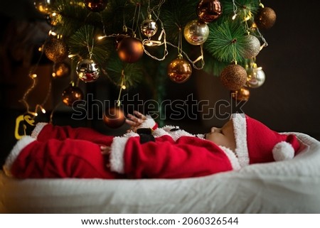 Closeup portrait of newborn baby. Cute Caucasian baby girl 4-5 months old in Santa costume lying on knitted cozy blanket in cocoon near decorated fir tree. Merry Christmas xmas and happy new year 2022