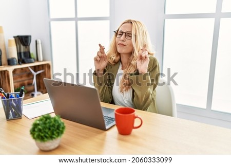 Beautiful blonde woman working at the office with laptop gesturing finger crossed smiling with hope and eyes closed. luck and superstitious concept. 
