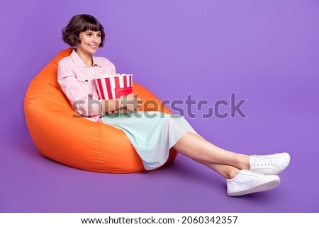 Full body photo of happy positive young woman sit chair watch film empty space pop corn isolated on violet color background