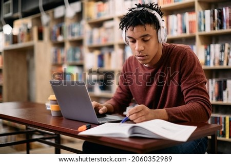 Young black student reading while using laptop and studying in a library. 