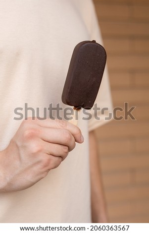 Junk food, summer, sweets and people concept - White male hands holding a chocolate ice cream cone on a stick on a background of a beige T-shirt and a brick wall outdoors with copy space