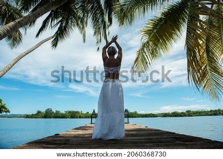 Beautiful blonde girl with short hair and in white tropical light and long dress standing on a pier in front of the lake surrounded by palm trees