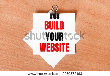 On a wooden table under a black paper clip lies a sheet of white paper with the text BUILD YOUR WEBSITE