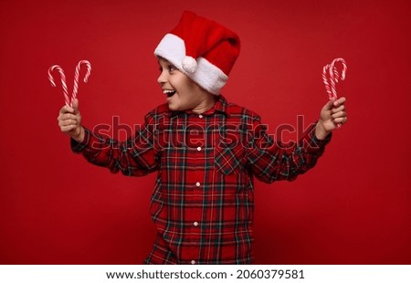 Beautiful child boy in checkered shirt and Santa Claus hat looking at Christmas lollipops, striped candy canes in his hands, isolated over red colored background with copy space for advertisement