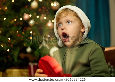 Cute boy in a Christmas hat opens a gift under a decorated Christmas tree. Happy boy with his mouth open in surprise holds a magic glowing box in his hands.