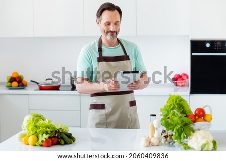 Photo of curious smart bearded man hold tablet read recipe wear kitchenware apron in kitchen apartment indoors