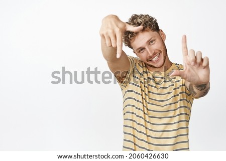 Happy blond caucasian man look through hand frames gesture and smiling, creating perfect pic, searching angle for photo, standing over white background