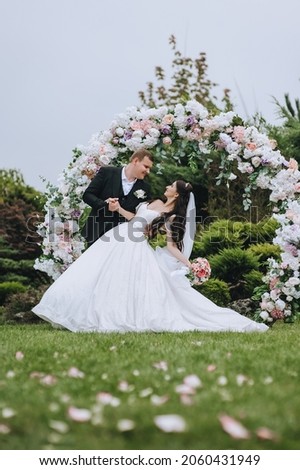 Stylish groom in a black suit and a bride in a white dress hug, dancing, holding hands at the ceremony, against the background of a round arch of flowers. Wedding portrait, photo of newlyweds.