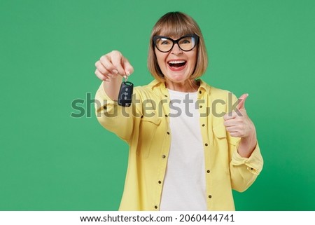 Elderly smiling happy caucasian woman 50s in glasses yellow shirt hold car keys fob keyless system show thumb up gesture isolated on plain green background studio portrait People lifestyle concept. Royalty-Free Stock Photo #2060444741