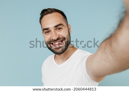 Close up young fun smiling happy caucasian man 20s wearing casual basic white t-shirt doing selfie shot pov on mobile phone camera isolated on plain pastel light blue color background studio portrait.